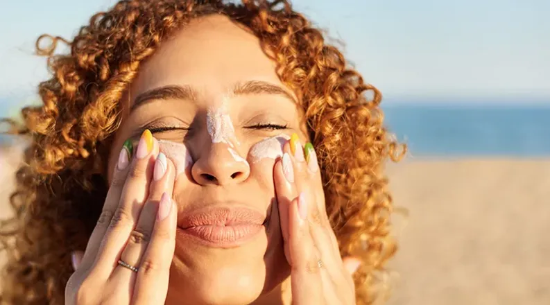 A woman with curly red hair applies sunscreen to her face wh