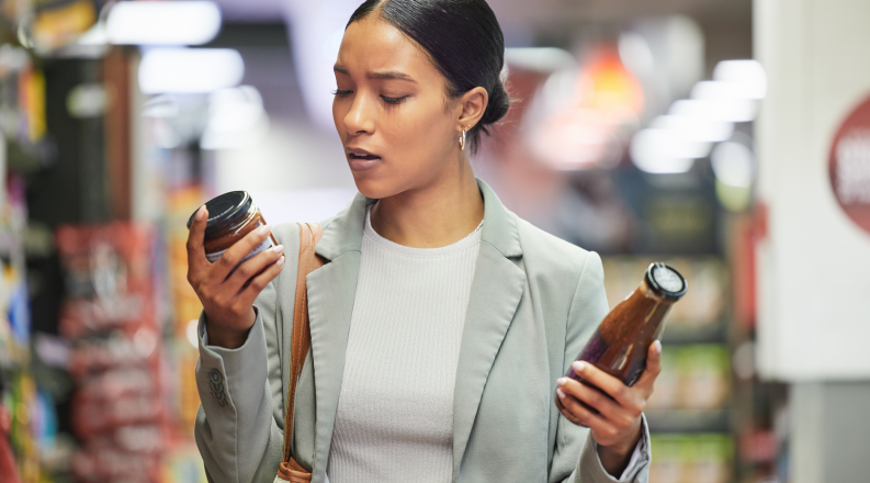 A woman in a grocery store holds a jar in each hand while reading a label on one product.