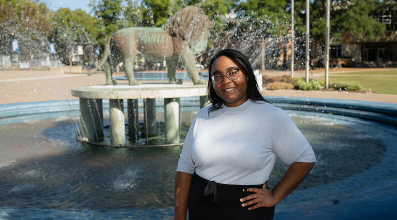 Destini Harrell, smiling at the camera, stands in front of the ODU lion fountain silhouetted against a blue sky.