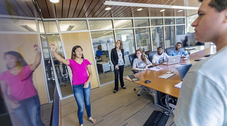 Students in conference room sit at table while looking at whiteboard.