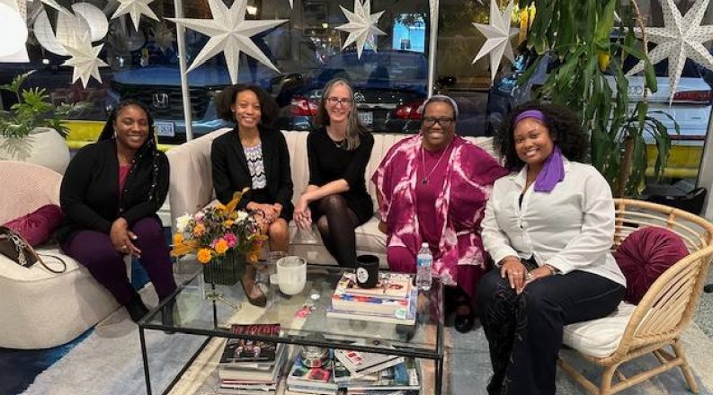 Five women sitting on chairs around a coffee table smile at the camera