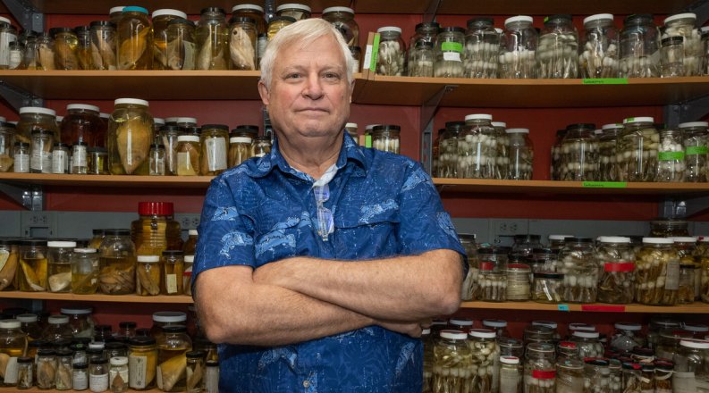 A man in a blue shirt poses in front of jars of biological samples.