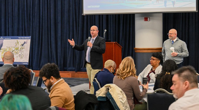 A man gestures as he presents in front of an audience.