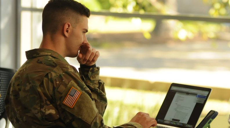 A student in military uniform works at a laptop.