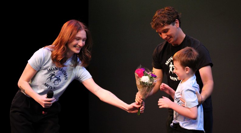 Baillie Ollila, a young woman with red hair wearing a Monarchs Move t-shirt, hands a young dance participant flowers, alongside another volunteer.