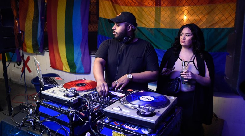 A DJ spins a record in front of a rainbow flag.