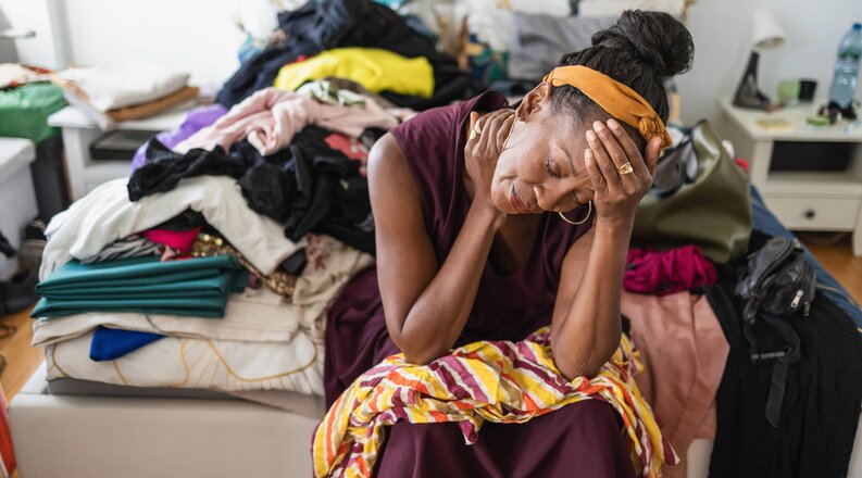 Overwhelmed woman  sitting on a messy bed covered with clothes with her head leaning on her hand.