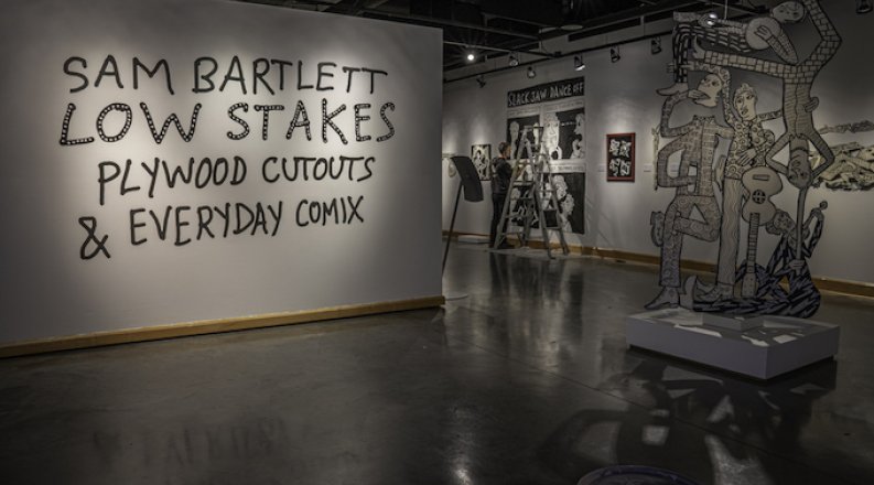 In the background, artist Sam Bartlett works on a comic panel for his exhibit, “Low Stakes: Everyday Comix and Plywood Cutouts” at the Baron and Ellin Gordon Art Galleries. Credit: Cullen B. Strawn