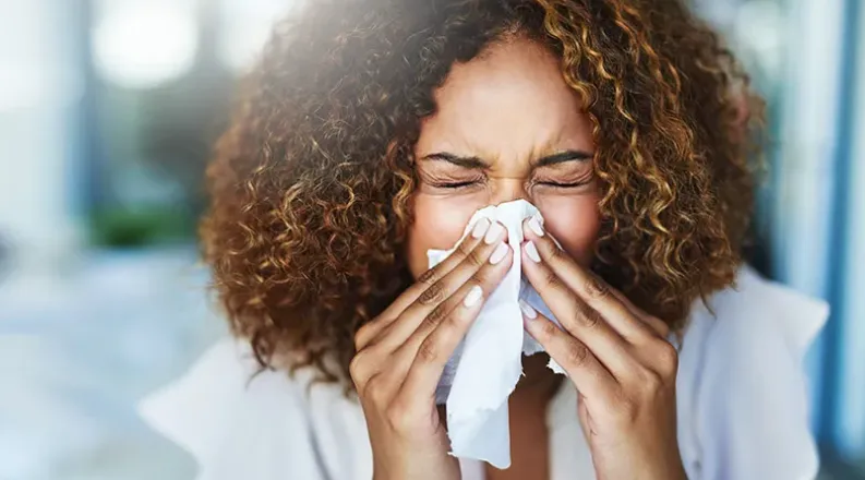 A woman with brown curly hair sneezes into a tissue.