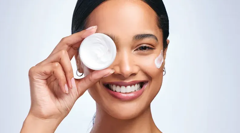 A smiling woman holds a small container of moisturizer as sh