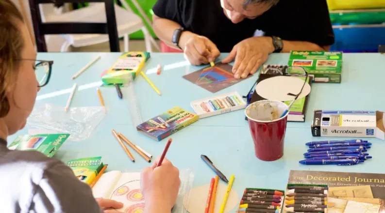 Individuals creating art at a table with various art supplie
