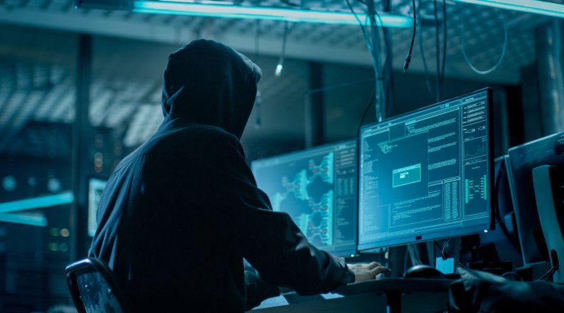 Shot from the Back to Hooded Hacker Breaking into Corporate Data Servers from His Underground Hideout. Place Has Dark Atmosphere, Multiple Displays, Cables Everywhere.