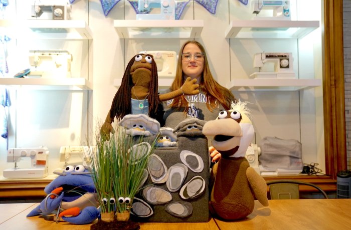 Sydney Walker poses with puppet characters from “The Amazing Neighborhood Mollusk Adventure” including Mr. Blue the crab, Reign the mermaid, a trio of oysters called the Reef Rascals and Esperanza, a brown pelican. Two clumps of marsh grass with googly eyes, named Tina and Flora, stand in the foreground.