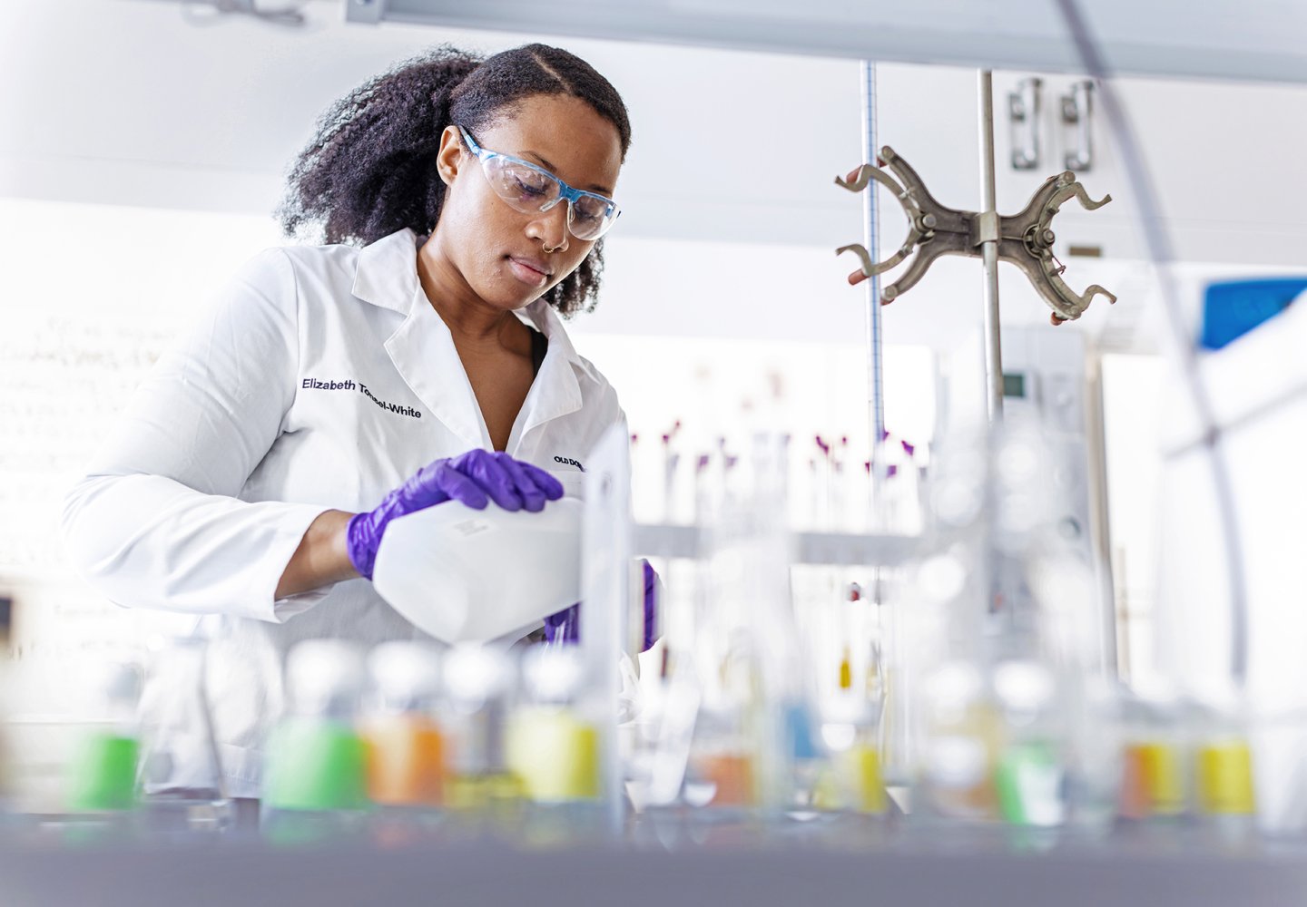 Student in a white lab coat and purple gloves in a chemistry lab.