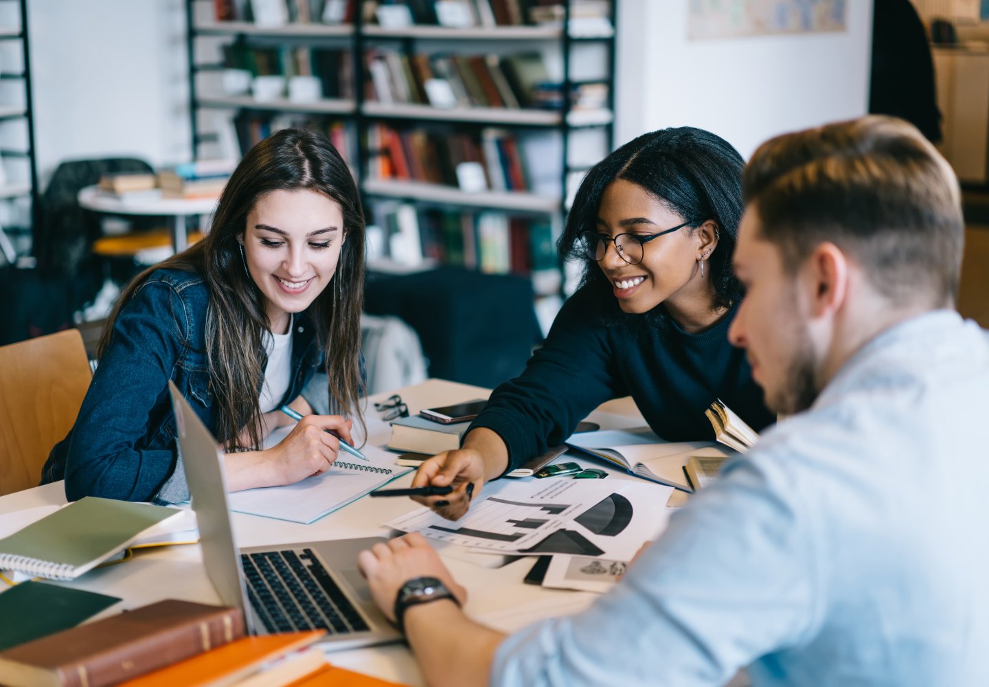 Three students watching educational video on laptop in library, skilled team of young people using netbook and wifi in library