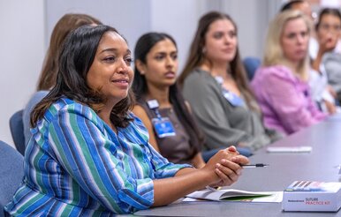 Students sit in row at table while looking on during presentation.