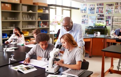 High School Students With Tutor Using Microscope In Biology 