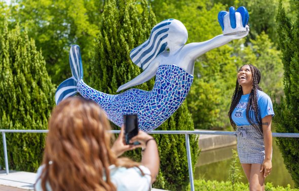 a student wearing an blue shirt is standing under the odu norfolk mermaid while another student takes her photo