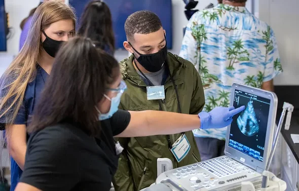Three students wearing masks look at an ultrasound monitor
