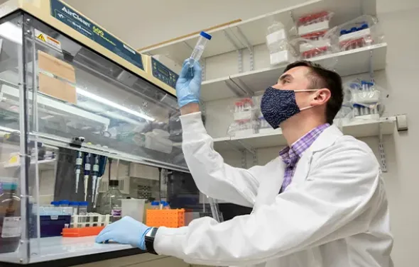A male student wearing a PPE mask, lab coat and gloves works