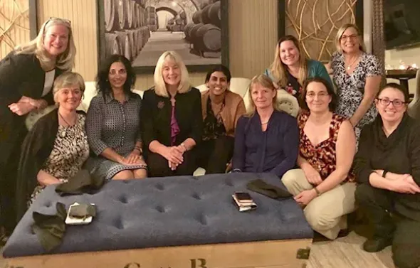 Ten women are gathered around an ottoman smiling at the came