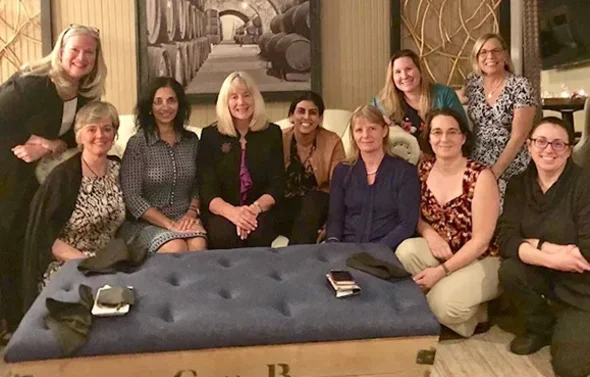 Ten women are gathered around an ottoman smiling at the came