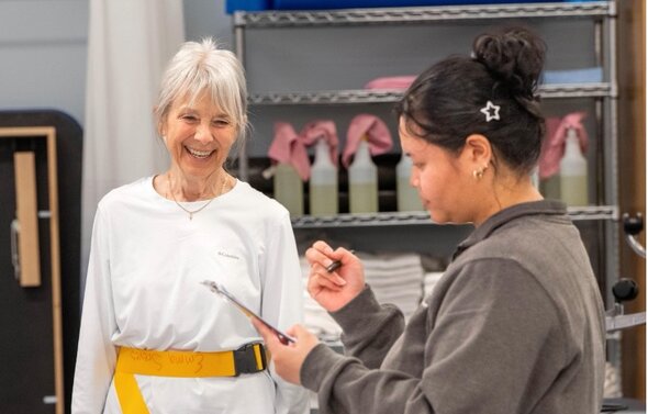 Female clinician writes on clipboard during physical therapy session with elderly woman.