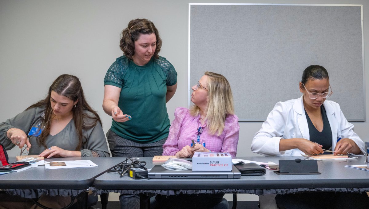 Nursing instructor talks to students sitting at table practicing with sutures.