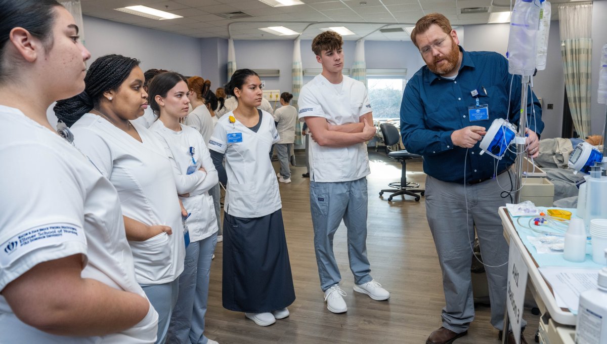 Group of nursing students watch as instructor demonstrates procedure in hospital setting.