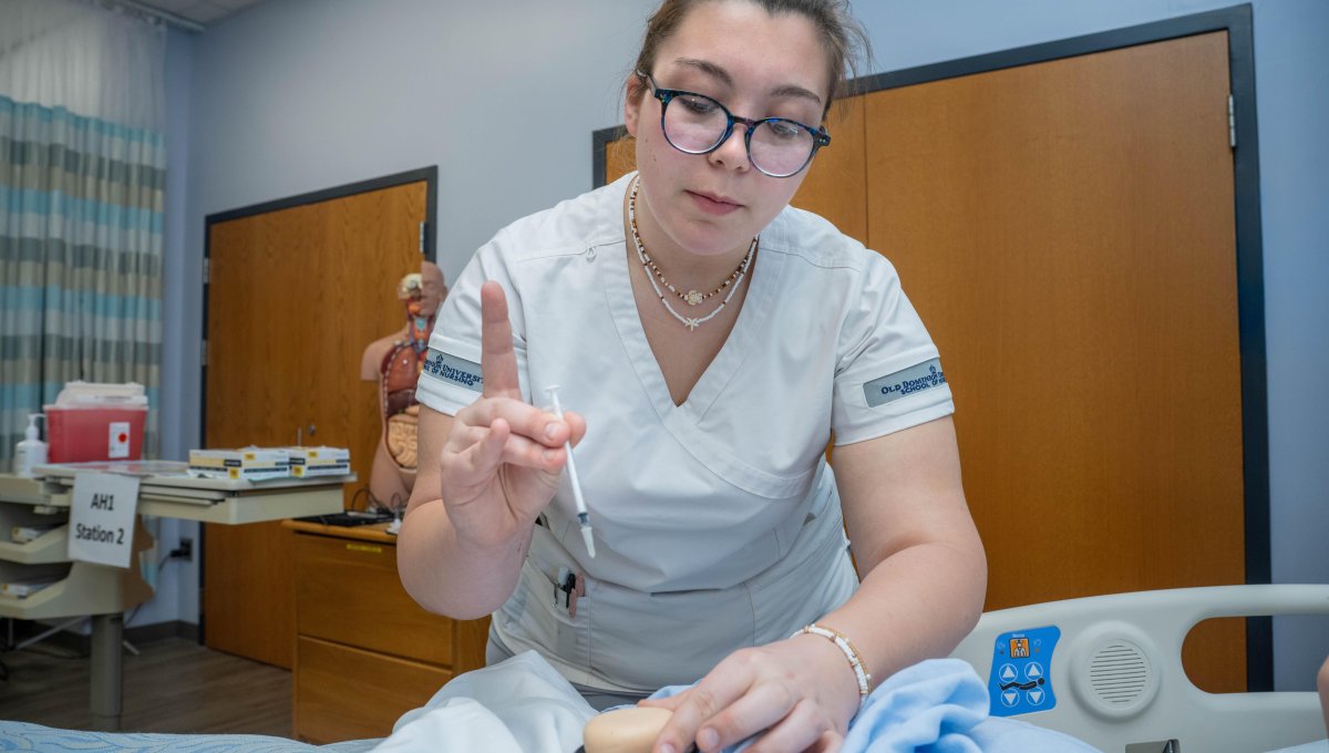Nursing student practices injecting needle into patient manikin.