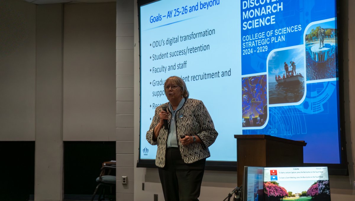 Dean Gail Dodge stands on stage holding a microphone welcoming 2025-2026 College of Sciences faculty and staff.