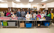 Officials and students from ODU and Norfolk Public Schools stand behind table in cafeteria kitchen with donated canned goods on top.