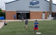 Young baseball players play a game in front of a building with a sign that says "ODU Monarchs: Paul A. Keyes Hitting Facility."