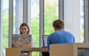 Two students sit at a table with their laptops open.