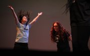  Two young women wearing Monarchs Move t-shirts stand on stage. One has her arms in the air, her hair flying upward as she dances.
