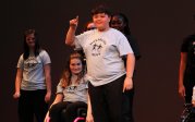 Several young participants in Monarchs Move t-shirts stand on stage. A boy in the front smiles as he holds up one finger.