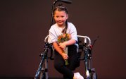 A young girl using a mobility walker holds flower and smiles on stage.