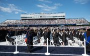 Students wearing academic robes and mortarboards stand in rows in a stadium.