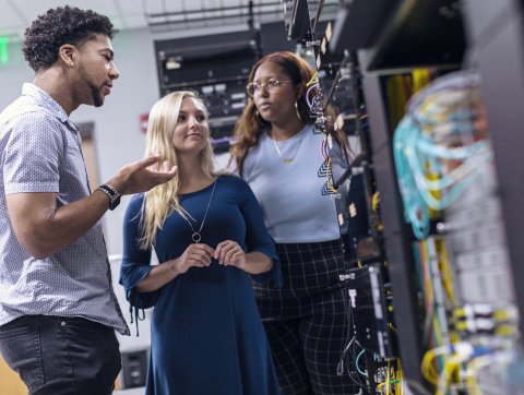 A group of three students are standing around electrical equipment in the cyber security lab
