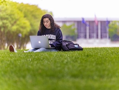A female student wearing an ODU sweatshirt is studying on Kaufman Mall with her laptop resting on her legs
