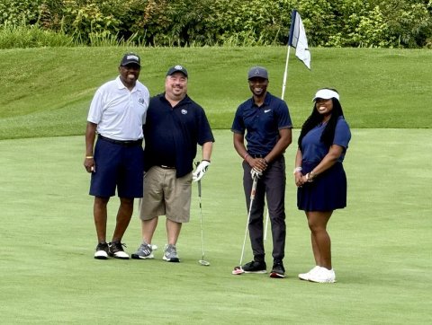 Four participants stand together on a golf course green, holding golf clubs and smiling for a group photo during an alumni golf outing.