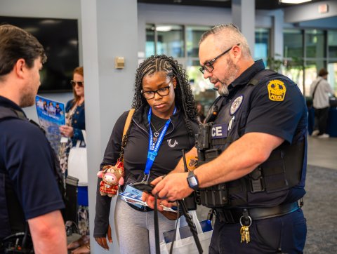 An Old Dominion University police officer in full uniform standing next to another officer and showing information on a mobile phone to a young female student wearing glasses and an ODU lanyard during a new student orientation event.