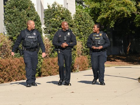 Three police officers in uniform walk down sidewalk while smiling.