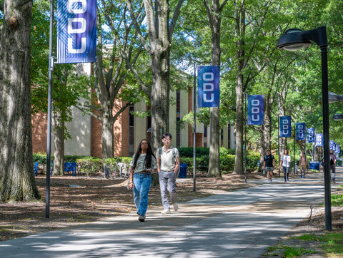 Students walk down campus sidewalk lined with trees and ODU flag poles.