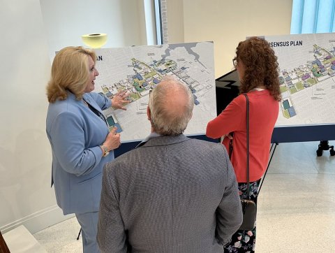 Three individuals review and discuss a campus development master plan displayed on presentation boards during a university planning event.