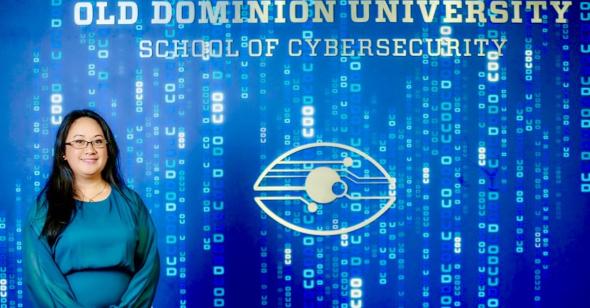 a dark haired professor stands in front of a wall with a cyber themed mural