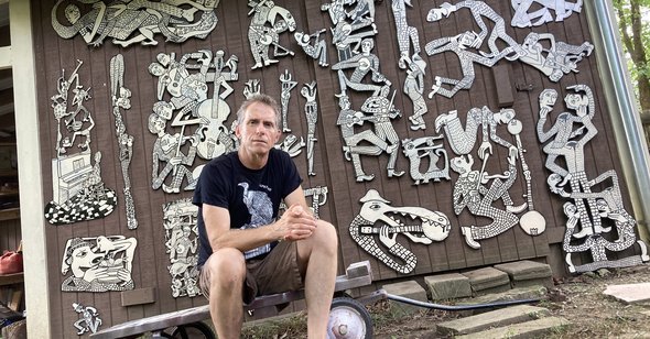 Man sits in front of building decorated with flat wooden sculptures.