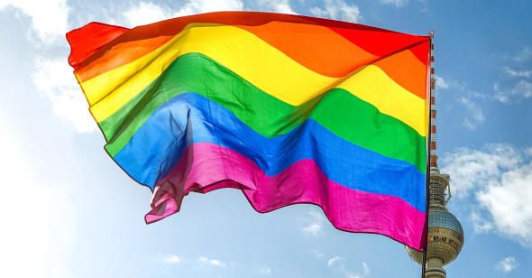 An LGBTQ pride flag waves against a backdrop of blue sky and white clouds.