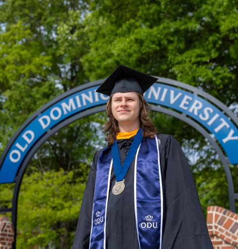 Alex Long, 2026 ODU Honors College graduate, smiles while wearing a cap and gown in front of an ODU campus sign.