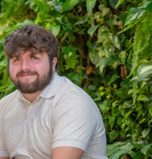 Adyn, a young White man with brown hair and beard wearing a beige collared shirt, sits against a background of greenery. 